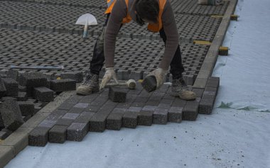 Paving taş işçisi bir şehir caddesinin inşaatı sırasında kaldırımları örülmemiş kumun üzerine yerleştiriyor ve yerine yerleştiriyor..