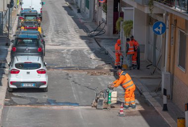 Modugno, İtalya - 20 Ekim 2025: Şehir caddesini tamir eden yüksek görüşlü turuncu üniformalı inşaat işçileri. Bir işçi fiber optik kablolar döşemek için asfalt kesme makinesi kullanıyor..
