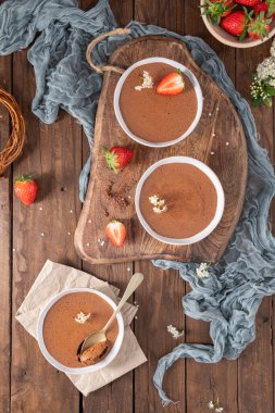 Three white bowls of creamy chocolate mousse, one with a golden spoon, are elegantly arranged on a wooden board and table with fresh strawberries and delicate white flowers