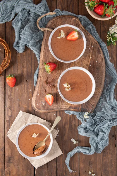 Three white bowls of creamy chocolate mousse, one with a golden spoon, are elegantly arranged on a wooden board and table with fresh strawberries and delicate white flowers