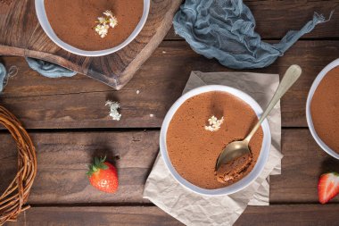 Üç beyaz kase yumuşak çikolatalı mus, biri altın kaşıkla taze çilekler ve narin beyaz çiçeklerle sanatsal bir şekilde ahşap bir masaya konur.