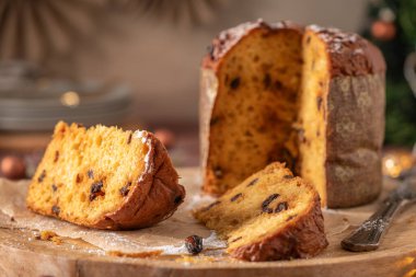 Altın Panettone dilimlerinin yakın çekimi ve arka planda tüm pasta, dağınık kuru üzümler ve yuvarlak ahşap bir kesme tahtasının üzerinde hafif bir toz şekeri içeren