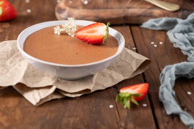 Beyaz bir kasedeki ipeksi çikolatalı mus, taze çilek dilimi ve küçük beyaz çiçeklerle süslenmiş, kırsal ahşap bir masanın üzerindeki buruşuk parşömen kağıdının üzerinde dinleniyor.