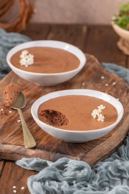 Beyaz kaselerde zengin çikolatalı mus tabakları, bir kaşık dolusu kaldırılmış, narin beyaz çiçeklerle süslenmiş, yumuşak mavi kumaşlı kırsal ahşap bir tahtanın üzerinde sunulmuş.