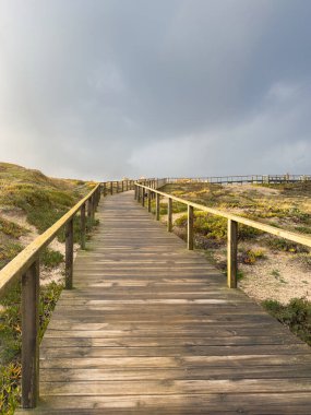 Uphill Wooden Boardwalk on Coastal Dunes Ovar A straight wooden boardwalk with railings ascends a sandy dune covered in low coastal vegetation under a dramatic, moody, gray sky