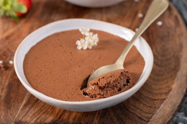 Beyaz bir kasede yumuşak çikolatalı mus, bir ısırığı kaldıran altın bir kaşık, küçük beyaz çiçeklerle süslenmiş, ahşap bir tahtanın üzerinde sunulmuş.