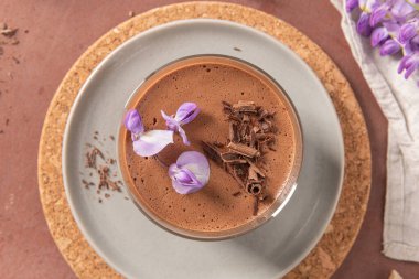 Çikolata parçacıkları ve mor çiçeklerle süslenmiş, tabakta şekillendirilmiş, bardaktaki kremalı çikolatalı mus ve mantar kaplama..