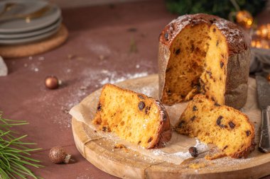 Koca bir Panettone, Noel süsleri, çam iğneleri ve kırmızımsı kahverengi bir masa arasında tahta bir tahtanın üzerinde oturan kuru üzüm ve şeker içeren iki parçayla kısmen dilimlenmiş.