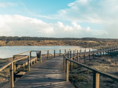 Ovar 'ın sahil lagünü kıyısındaki tahta patikanın kıyısında tahta kaldırım yolu sakin bir kıyı lagününden geçerek Furadouro Sahili' ndeki dramatik bulutlu mavi gökyüzü Ovar Portekiz 'de yoğun bir çam ormanına doğru ilerliyor.