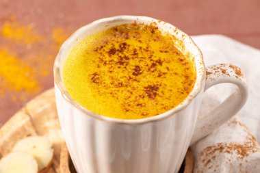 Sıcak seramik bir kupa köpüklü altın zerdeçal latte ile kaplanmış, mantar altlığı üzerinde yumuşak bir terracotta arka planına yaslanmış baharat tozuyla.