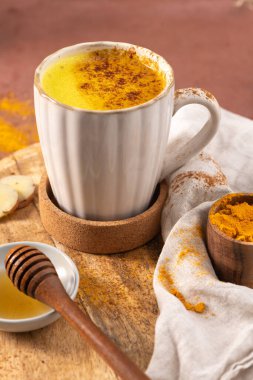 Taze zencefil kökü zerdeçal tozu tarçın ve sıcak bir bütünsellik içeceği için bal ile çevrili beyaz kurdeleli fincanda canlı altın sütlü latte. Baharatlı Sağlıklı Altın Süt Sütlü Süt