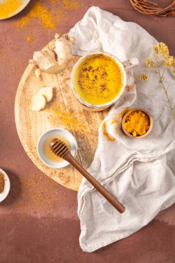 Taze zencefil, bal ve ahşap bir tahtanın üzerinde sade keten kumaştan yapılmış zerdeçal tozuyla çevrili altın sütlü lattenin yukarıdan aşağı görüntüsü.