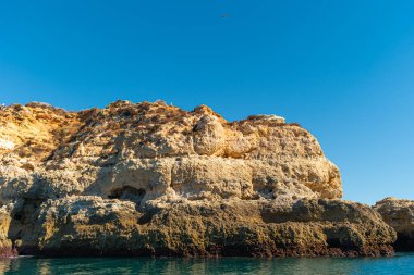 Portekiz 'in Algarve bölgesindeki berrak mavi gökyüzünün altında, sakin zümrüt Atlantik sularının üzerinde engebeli altın kireçtaşı uçurumlar kulesi.
