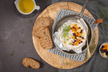 Yukarıdan bakıldığında, kurutulmuş meyve ve fındıkla süslenmiş kaymak gibi fırınlanmış bir peyniri, rustik ahşap bir tabakta ızgara çavdar ekmeği ve zeytin yağıyla servis edilmiş ve koyu masa ile süslenmiş bir kremalı Camembert peyniri yer alıyor.