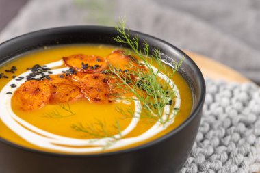 Kızarmış havuç dilimleri, krem kıvrımları ve kırsal gri bir zemin üzerinde taze dereotları olan siyah bir kasede servis edilen yumuşak havuç çorbası.