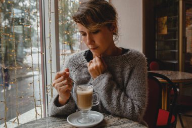 Sıcak ceketli, kahverengi saçlı genç bir kadın bir kafede, pencerenin yanındaki masada oturuyor ve kahveyi karıştırıyor.