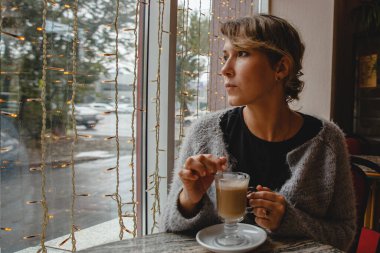 Sıcak ceketli, kahverengi saçlı genç bir kadın bir kafede pencerenin yanındaki masada oturuyor ve kahveyi karıştırıyor. Yüksek kalite fotoğraf