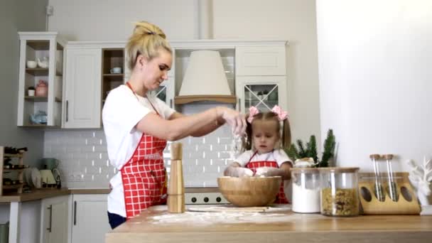 En ung blond mamma med en liten dotter i förkläden lagar mat vid bordet i köket.. — Stockvideo