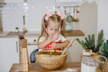 Beyaz tişörtlü, kırmızı önlüklü dört yaşında sarışın bir kız, mutfakta ahşap bir masada hamur yoğuruyor.