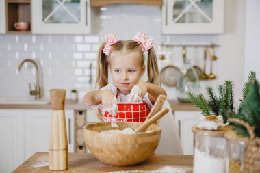 Beyaz tişörtlü, kırmızı önlüklü dört yaşında sarışın bir kız, mutfakta ahşap bir masada hamur yoğuruyor.