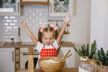Beyaz tişörtlü, kırmızı önlüklü dört yaşında sarışın bir kız, mutfakta ahşap bir masada hamur yoğuruyor.