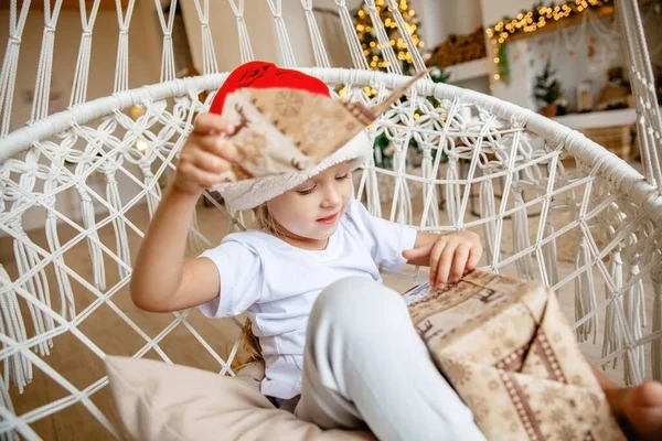Чотирирічна дівчинка у капелюсі Санти сидить у хиткому кріслі на задньому плані ялинки і відкриває подарунок. — стокове фото