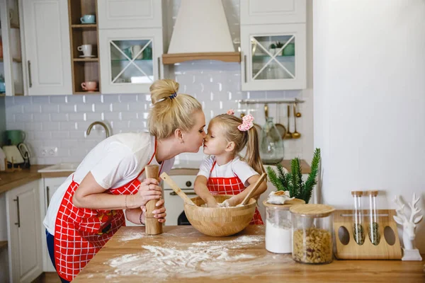 Ξανθιά μαμά με κόκκινη ποδιά φιλάει την κόρη της κατά την προετοιμασία της ζύμης στην Χριστουγεννιάτικη κουζίνα στο σπίτι — Φωτογραφία Αρχείου