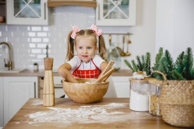 Beyaz tişörtlü, kırmızı önlüklü dört yaşında sarışın bir kız, mutfakta ahşap bir masada hamur yoğuruyor.