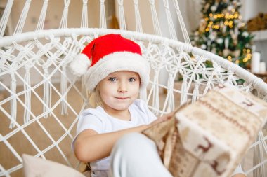 Dört yaşında Noel Baba şapkalı bir kız hasır bir sandalyede oturuyor ve bir Noel ağacının arka planında hediyesini açıyor.