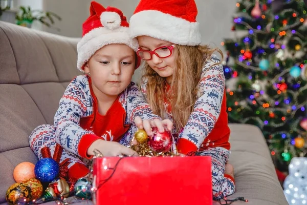 Irmãozinho e irmã em chapéus de Papai Noel no sofá contra a árvore de Natal tirar decorações da caixa — Fotografia de Stock