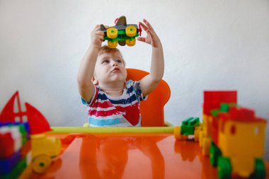 Küçük sarışın bir çocuk masasında parlak plastik oyuncakların arasında oturuyor ve uçakla oynuyor.