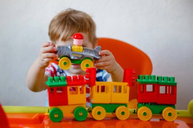 Evde turuncu bir çocuk masasında oturan küçük sarışın bir çocuk renkli plastik bir inşaat setiyle oynuyor.