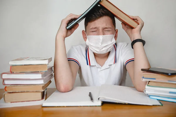 A teenager in a mask at home sitting at lessons covers his head with textbooks. Coronavirus concept. High quality photo