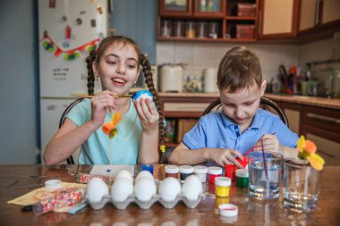 Esmer erkek ve kız kardeş Paskalya yumurtalarını evde, mutfakta masada oturarak süslerler..
