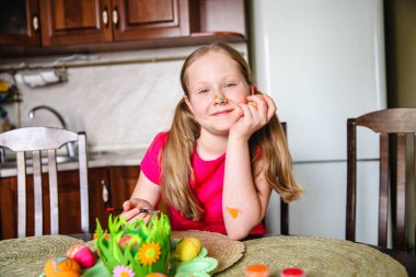 Uzun sarı at kuyruklu ve boyalı burunlu bir kız dekore edilmiş Paskalya yumurtalarının önünde mutfak masasında oturuyor. Yüksek kalite fotoğraf