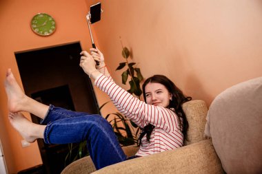 Genç bir kız sandalyede oturuyor ve akıllı telefonuyla selfie çubuğu tutuyor. Evdeki internet iletişimi. Yüksek kalite fotoğraf