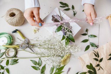 Bir çiçekçi hediye kutusunu çiçeklerle süslüyor ve beyaz bir masaüstünde kurdele takıyor. Üst görüntü. Sadece eller çerçevede.