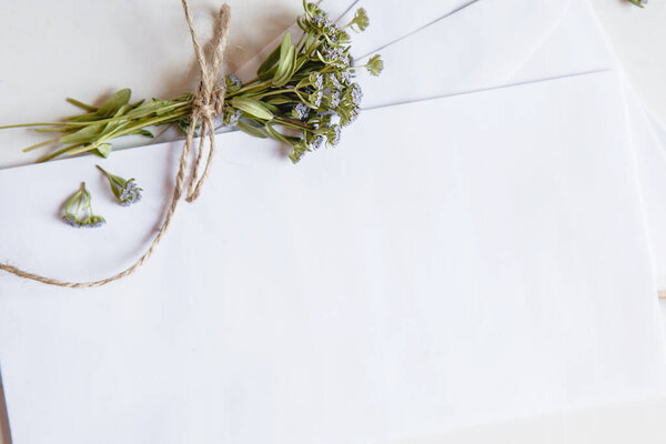  A white blank envelopes with a small bouquet of wildflowers tied with twine on a white wooden background. View from above