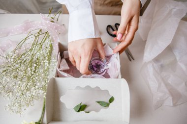 Bir bayan dekoratör beyaz bir hediye kutusuna dekoratif pembe bir mum koyar. Masanın yanında çiçekler ve çiçekçilerin aletleri var. Üst Manzara. Yüksek kalite fotoğraf