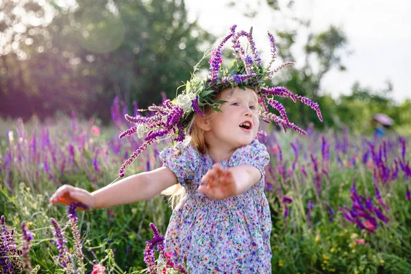 Una piccola ragazza bionda felice in una ghirlanda di salvia e un vestito colorato in un campo fiorito estivo — Foto Stock