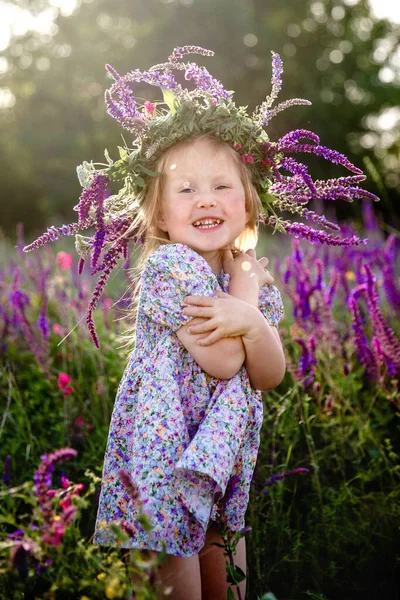 Una piccola ragazza bionda felice in una ghirlanda di salvia e un vestito colorato in un campo fiorito estivo — Foto Stock