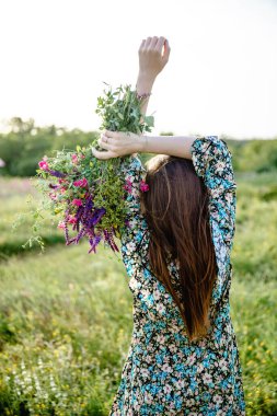 Renkli kısa elbiseli, uzun saçlı genç bir kadın kameraya arkası dönükken kaldırılmış ellerinde bir buket yabani çiçek tutuyor.