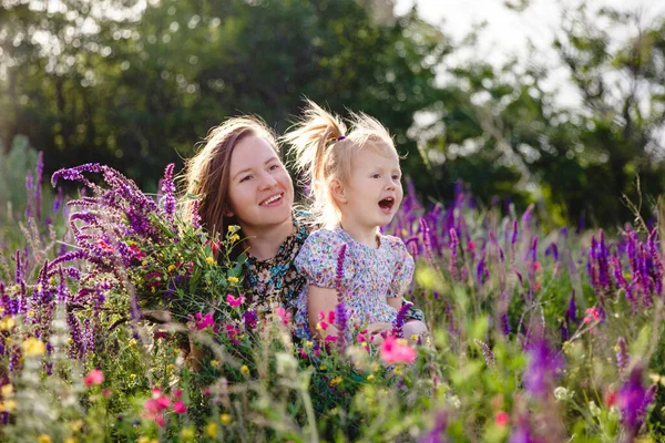 Счастливые блондинка мама и дочь в летнем цветущем поле с букетом диких цветов в руках — стоковое фото