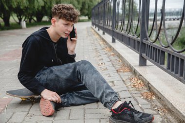 Avrupalı kıvırcık saçlı, siyah kapüşonlu bir genç kaykaylı bir sokakta oturuyor ve telefonda konuşuyor.