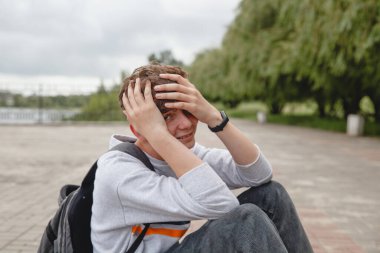 Kıvırcık saçlı, gri kazaklı, kot pantolonlu ve sırt çantalı bir Avrupalı genç kaldırımda oturuyor ve elleriyle yüzünü örtüyor.