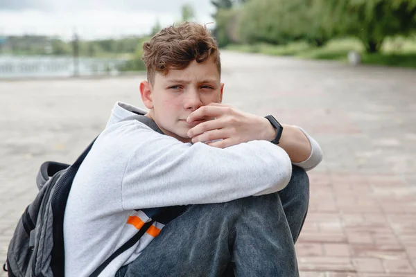 A curly-haired, thoughtful European teen in a gray jumper, jeans with a satchel on his back is sitting on the pavement — Stock Photo, Image