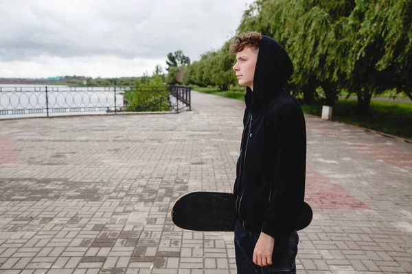 Um adolescente de cabelos encaracolados europeu em um capuz preto com um skate em suas mãos fica no dique. Vista do perfil — Fotografia de Stock