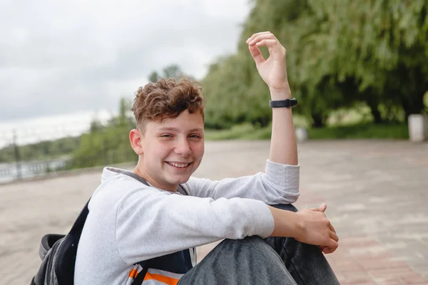 Ein gelockter lachender europäischer Teenager in grauem Pullover, Jeans und mit Schulranzen auf dem Rücken sitzt auf dem Bürgersteig. — Stockfoto