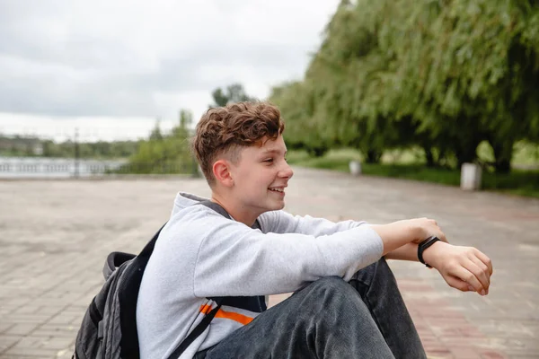 Un adolescente europeo dai capelli ricci ridente in un maglione grigio, jeans e con una borsa sulla schiena è seduto sul marciapiede — Foto Stock