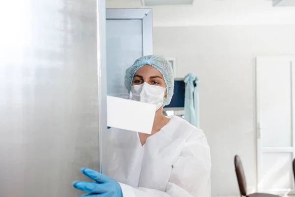 Una joven enfermera en traje de protección, gorra, máscara y guantes toma documentos de un casillero en el consultorio médico — Foto de Stock